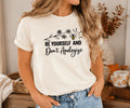 A woman wearing a cream-colored graphic t-shirt with a black printed design featuring a bee and the text 'BE YOURSELF AND Don't Apologize', paired with blue jeans in a cozy indoor setting.