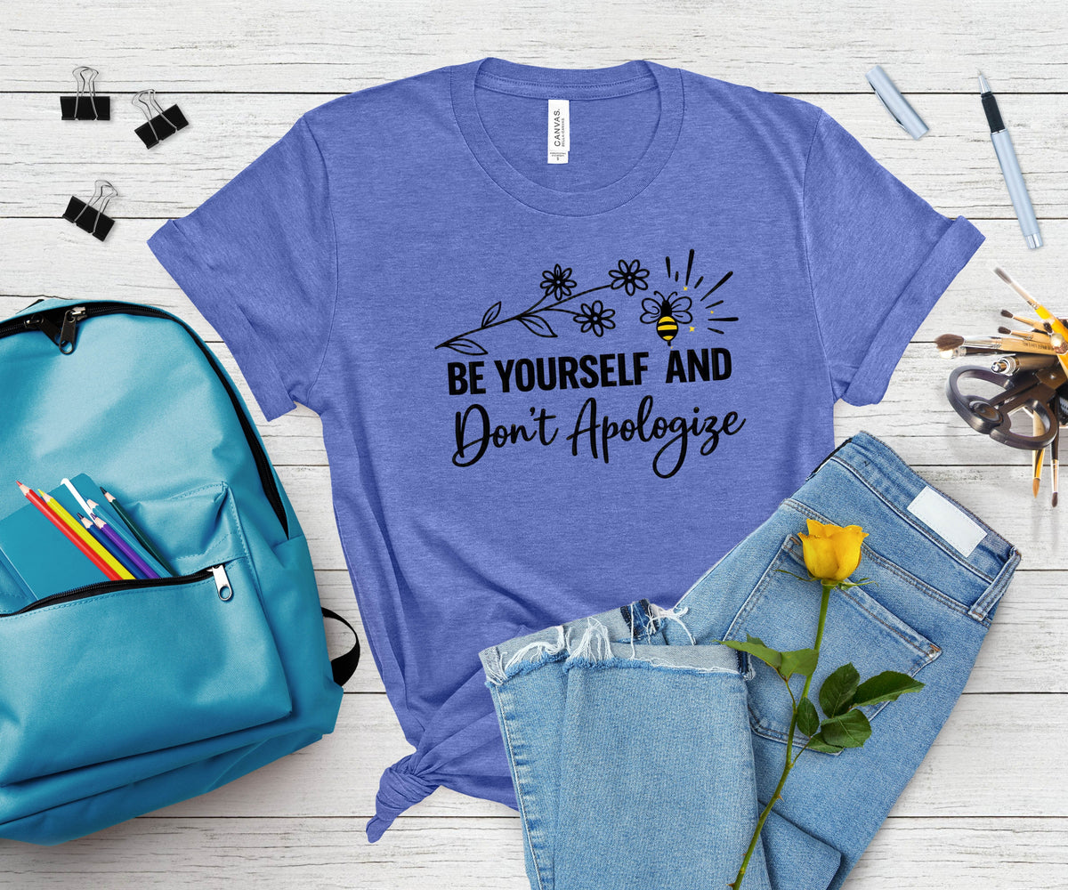 A heathered periwinkle blue graphic t-shirt with a black printed design that reads 'BE YOURSELF AND Don't Apologize' with a bee and flowers. The shirt is styled in a flat-lay arrangement on a white wood background, surrounded by a teal backpack, light-wash jeans, a yellow rose, and office supplies.