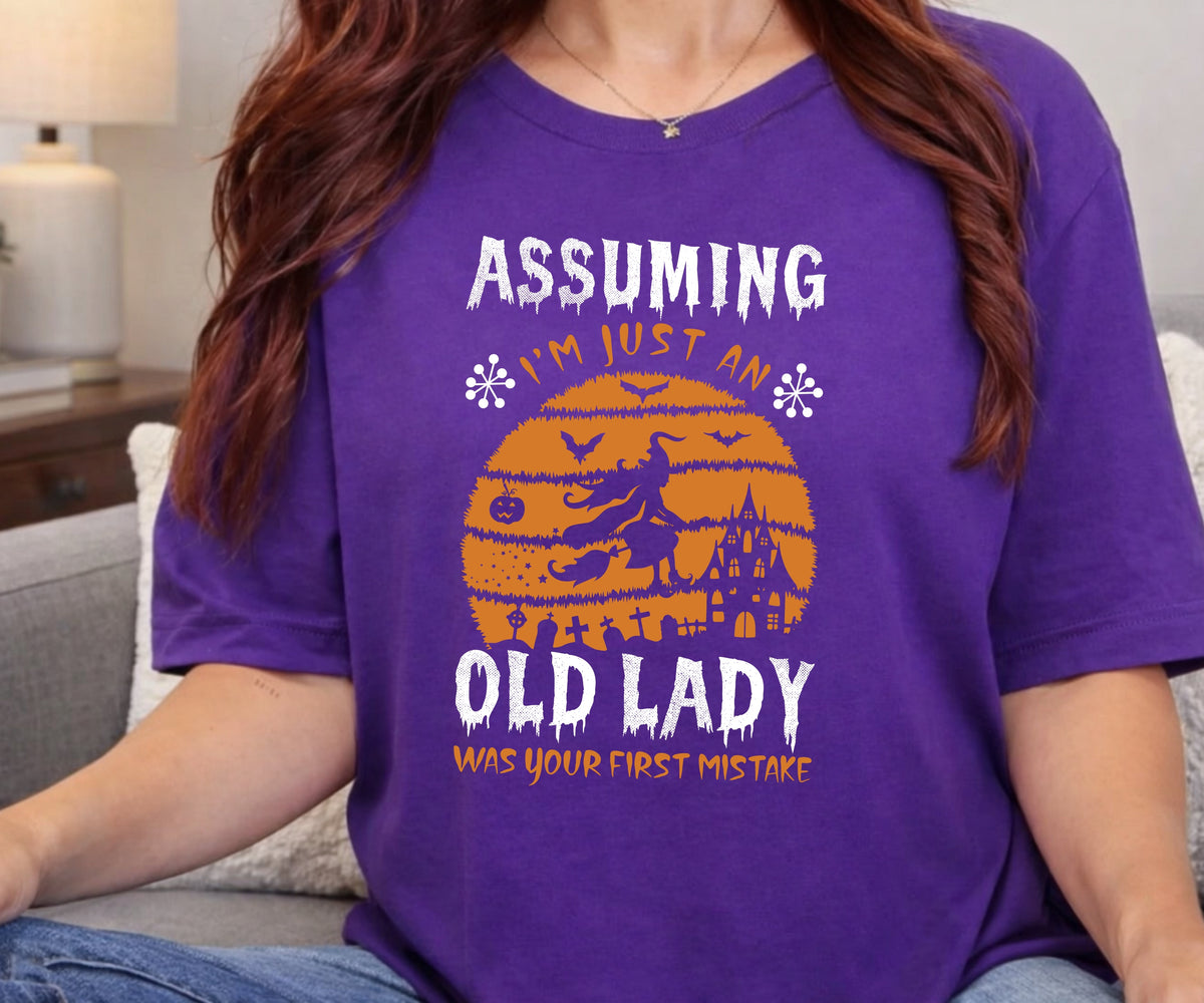 A woman wearing a purple graphic t-shirt with a Halloween-themed design that reads 'ASSUMING I'M JUST AN OLD LADY WAS YOUR FIRST MISTAKE' in white and orange text over a witch riding a broomstick silhouette.