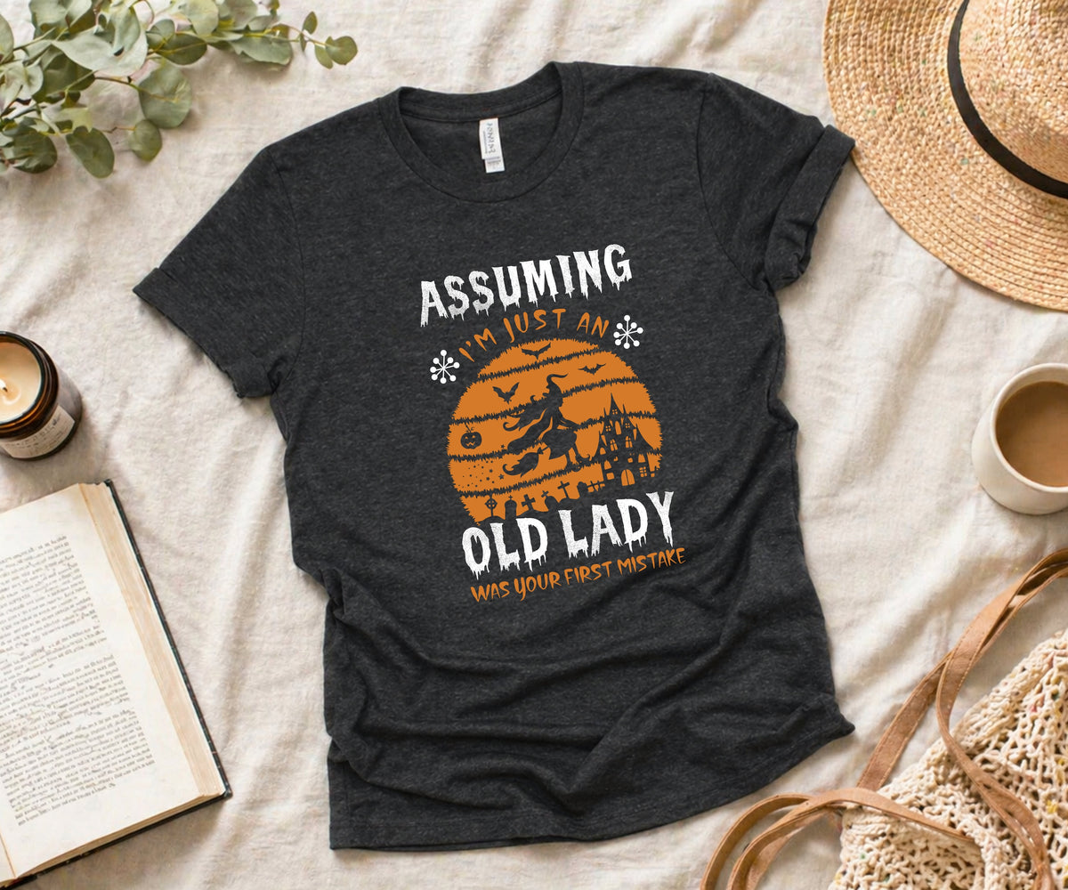 A dark heather gray graphic t-shirt laid flat on a beige fabric surface, surrounded by lifestyle props including a straw hat, a book, a candle, a cup of coffee, and eucalyptus leaves. The t-shirt features a retro orange graphic of a witch riding a broomstick in front of a sunset, with the text 'ASSUMING I'M JUST AN OLD LADY WHO'S YOUR FIRST DATE'.