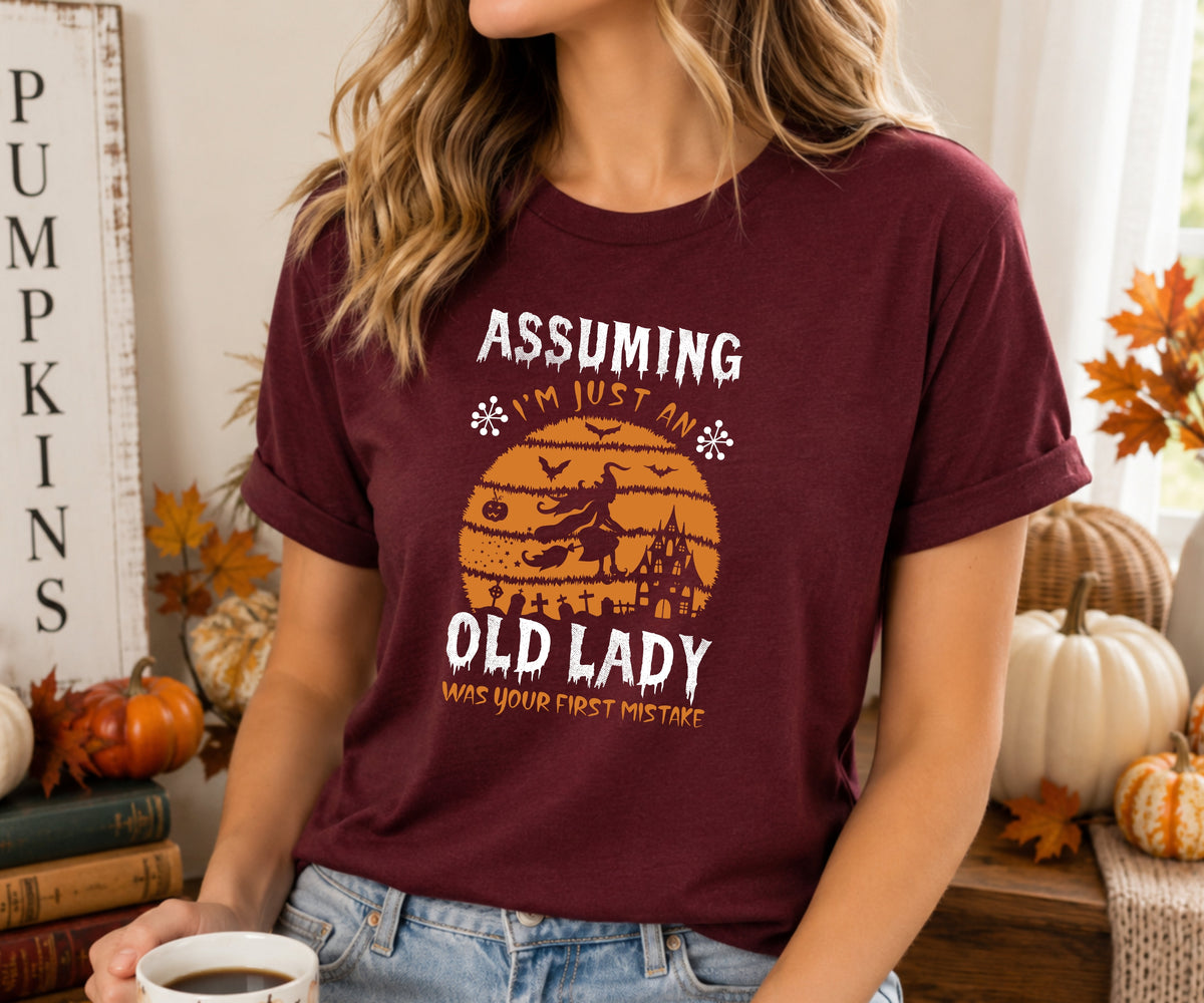 A woman wearing a maroon short-sleeve graphic t-shirt with a Halloween-themed print that reads 'ASSUMING I'M JUST AN OLD LADY WAS YOUR FIRST MISTAKE' in white and orange. The background is a cozy, autumnal setting with pumpkins, fall leaves, and a rustic sign.