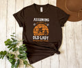 A dark brown graphic t-shirt laid flat on a white wood plank background, styled with a brown hat, green eucalyptus leaves, and a knot at the bottom hem. The t-shirt features a retro orange and white graphic with the text 'ASSUMING I'M JUST AN OLD LADY WAY YOUR FIRST MISTAKE'.