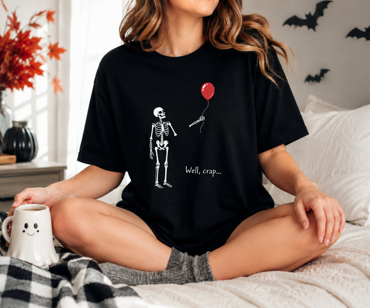 A woman sitting cross-legged on a bed wearing a black graphic t-shirt with a white skeleton holding a red balloon and the text 'Well, crap.