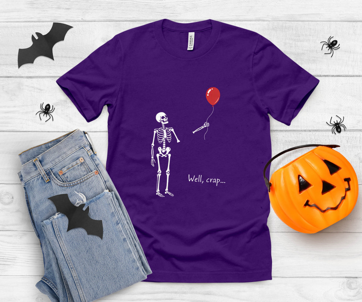 A purple short-sleeve graphic T-shirt laid flat on a white wood background, styled with jeans, a jack-o'-lantern bucket, and black bat and spider cutouts. The shirt graphic shows a white skeleton holding a red balloon with the text 'Well, crap.