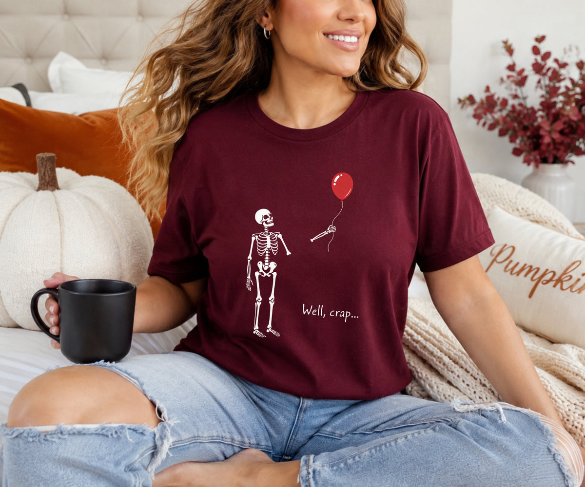 A woman sitting on a bed wearing a maroon graphic t-shirt with a white skeleton and a red balloon, holding a black mug. The background features a cozy bedroom setting with a white pumpkin, a cream tufted headboard, and a beige knit blanket.