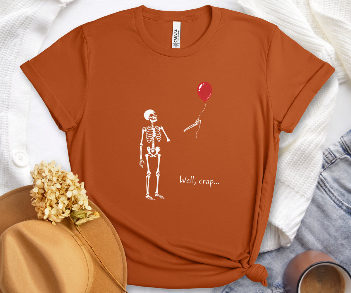 An orange short-sleeve graphic T-shirt laid flat on a white textured blanket with a tan hat, dried flowers, and denim jeans around it. The shirt has a white skeleton drawing reaching for a red balloon with the text 'Well, crap.
