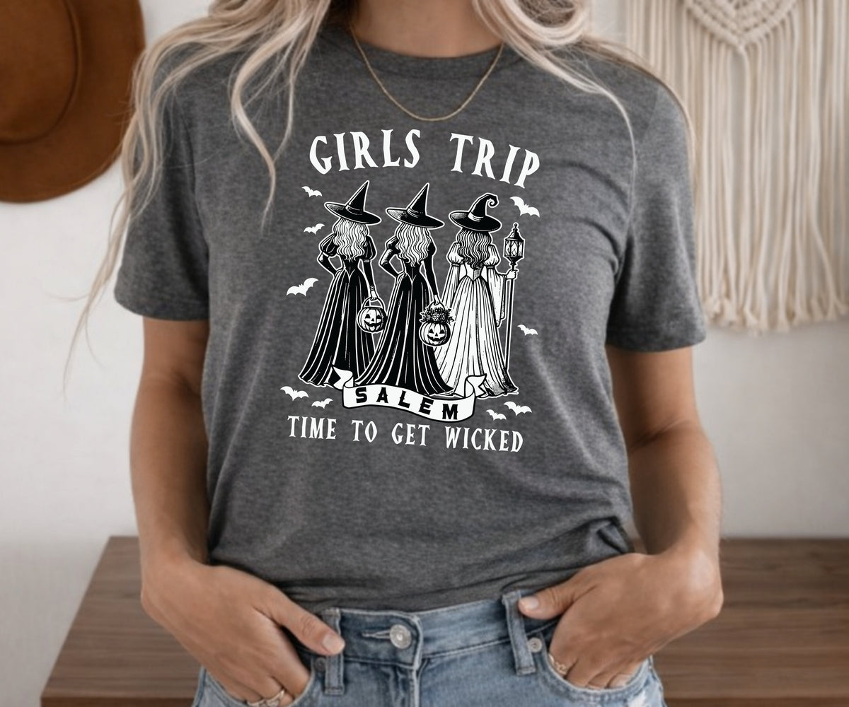 A woman wearing a dark heather gray graphic t-shirt with a white print of three witches and the text 'GIRLS TRIP SALEM TIME TO GET WICKED'.