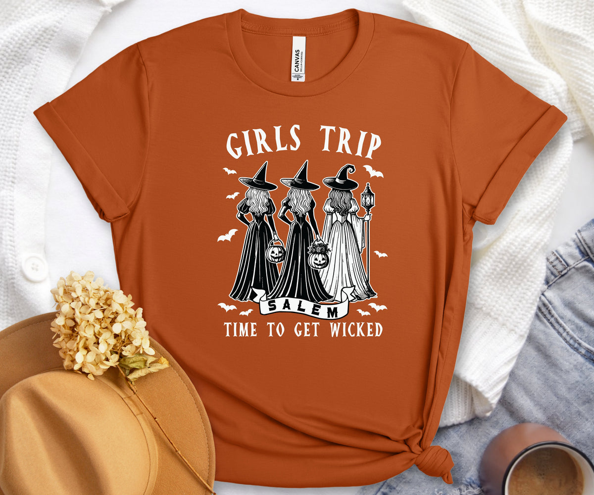 An orange short-sleeve graphic T-shirt laid flat on a white textured blanket, styled with a tan hat, dried flowers, denim, and a small cup. The shirt has a black-and-white witch illustration and the text 'GIRLS TRIP SALEM TIME TO GET WICKED' across the front.