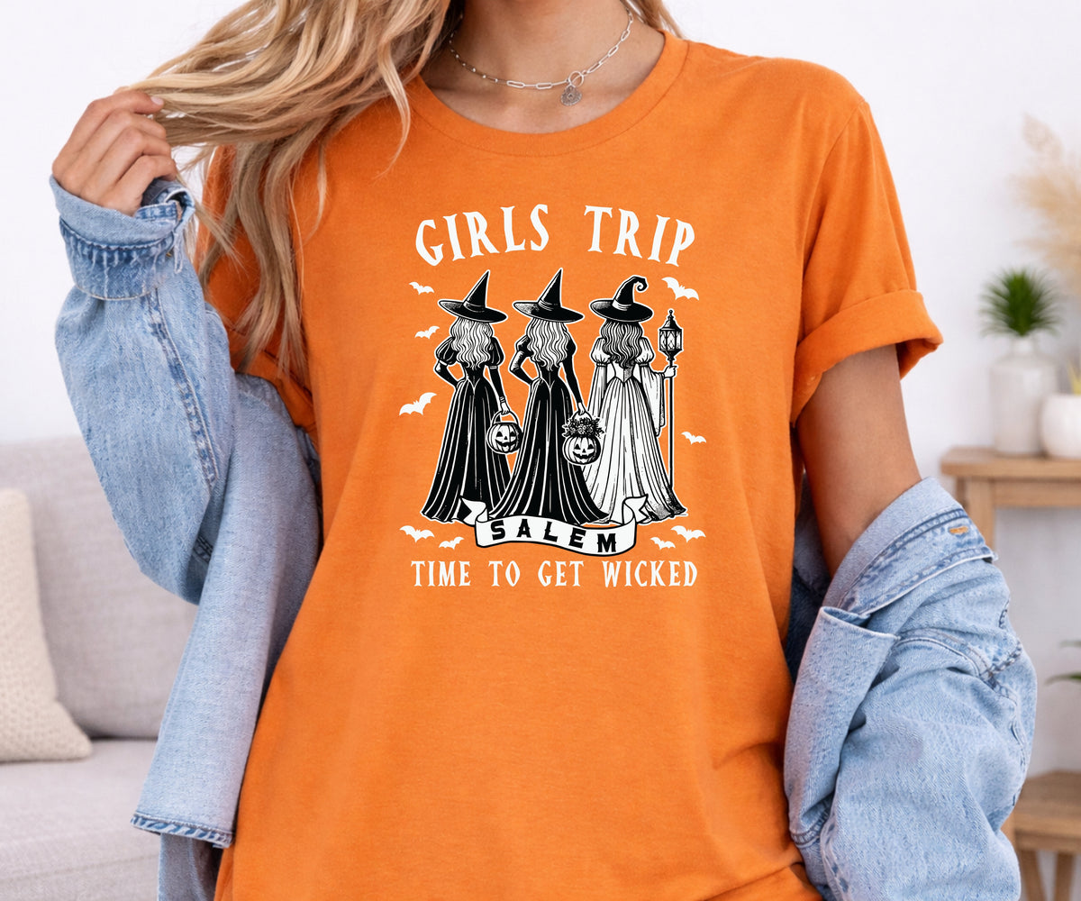 A woman wearing an orange graphic t-shirt with a Halloween design featuring three witches and the text 'GIRLS TRIP Balm TIME TO GET WICKED', styled with a light blue denim jacket draped off her shoulders.