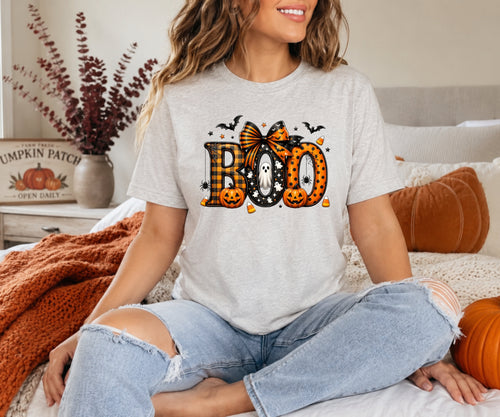 A woman sitting cross-legged on a bed wearing a light gray heather t-shirt with a Halloween graphic that reads 'BOO' in orange jack-o'-lantern patterns, black bats, and a witch hat. She is wearing light-wash ripped jeans in a cozy bedroom setting with autumn decor.