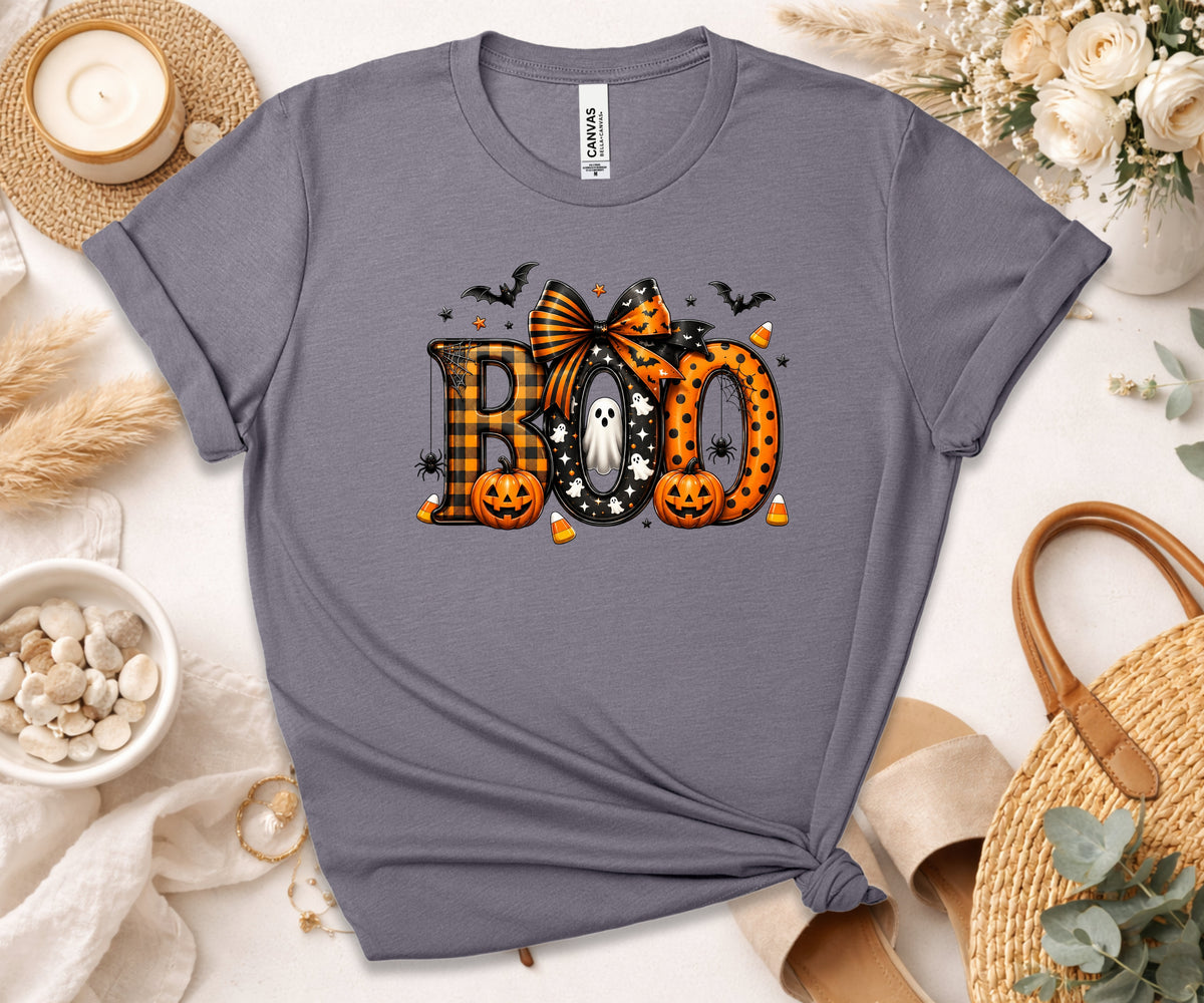 A slate gray short-sleeve t-shirt with a Halloween graphic reading 'BOO' in plaid and orange polka-dot lettering, styled in a flat lay with a candle, flowers, and woven accessories.