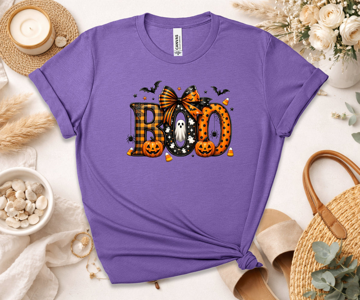 A purple short-sleeve t-shirt with a 'BOO' Halloween graphic, styled in a flat lay with a candle, flowers, and straw sandals.