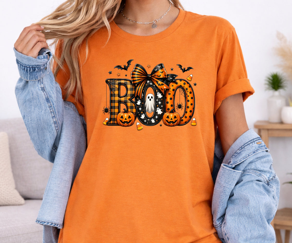 A woman wearing a bright orange graphic t-shirt with a Halloween 'BOO' design, styled with a light blue denim jacket draped off her shoulders in a cozy indoor setting.