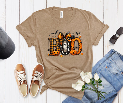 A tan heather short-sleeve graphic T-shirt laid flat on a white wood background, styled with a pair of blue jeans, white roses, tan sneakers, sunglasses, and a dark cap. The shirt has a large Halloween-themed 'BOO' graphic in orange, black, white, and gray with bats and pumpkins.