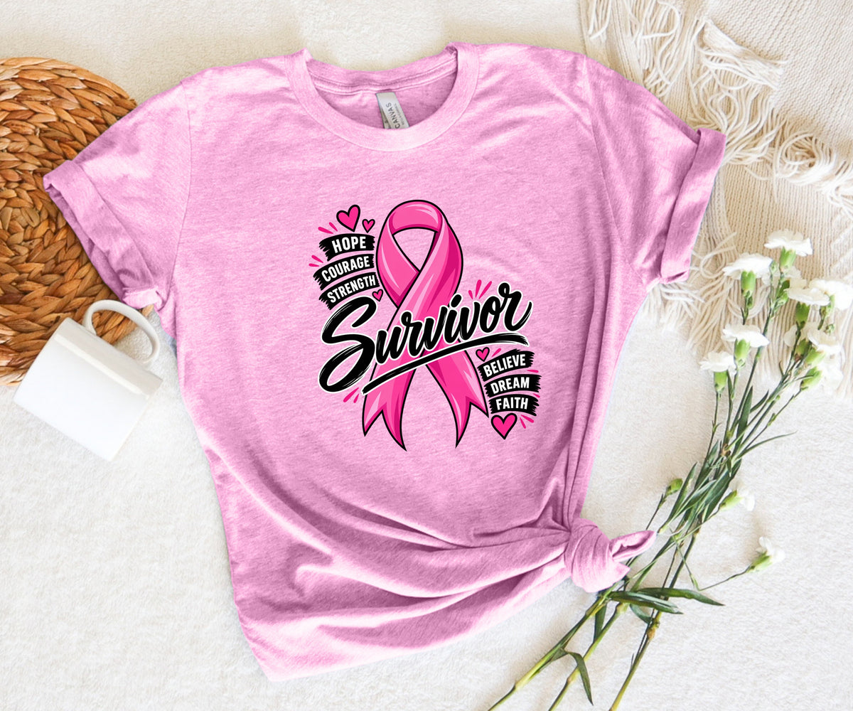 A pink short-sleeve graphic T-shirt laid flat on a white textured surface, styled with a woven mat, white mug, and small white flowers. The shirt has a large pink awareness ribbon and the word 'Survivor' with smaller text reading 'Hope Courage Strength Love Believe Dream Faith'.