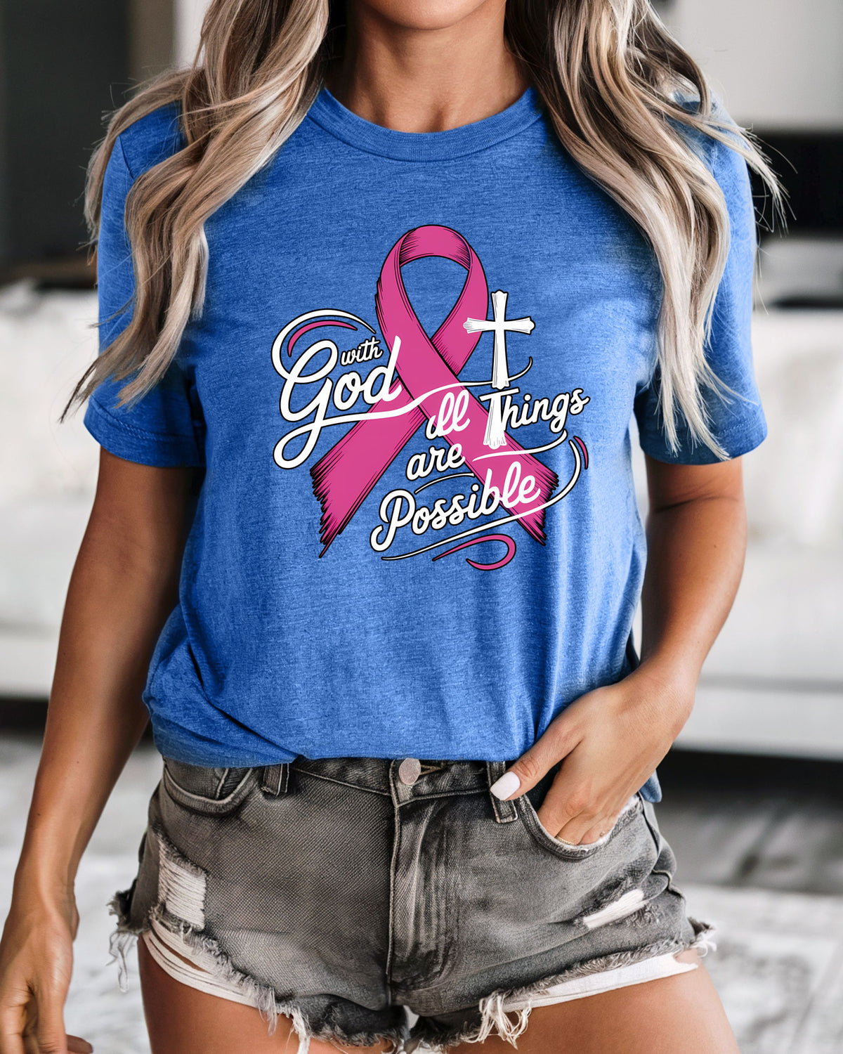 A woman wearing a heather blue graphic t-shirt with a pink ribbon and the text 'With God all Things are Possible' in white and pink, paired with distressed denim shorts.