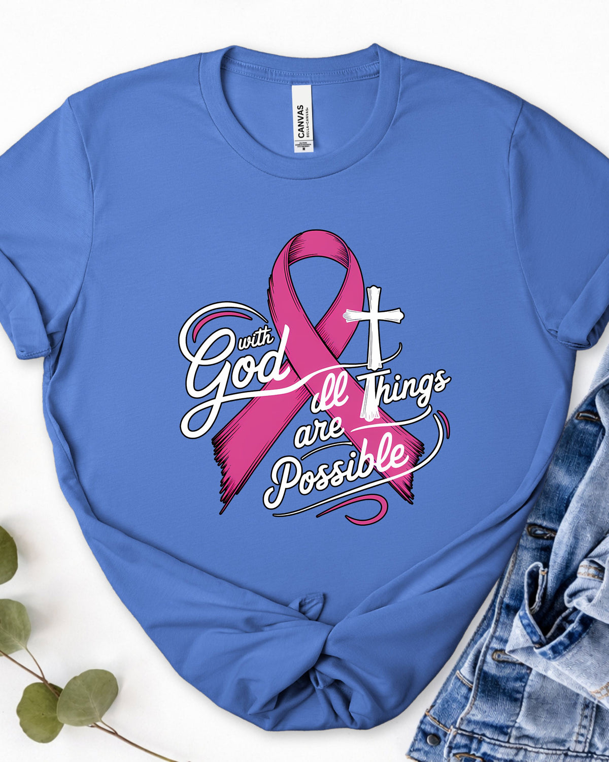 A royal blue short-sleeve t-shirt with a pink and white graphic reading 'with God all Things are Possible' alongside a pink awareness ribbon and white cross, styled flat-lay with denim shorts and green leaves on a white background.