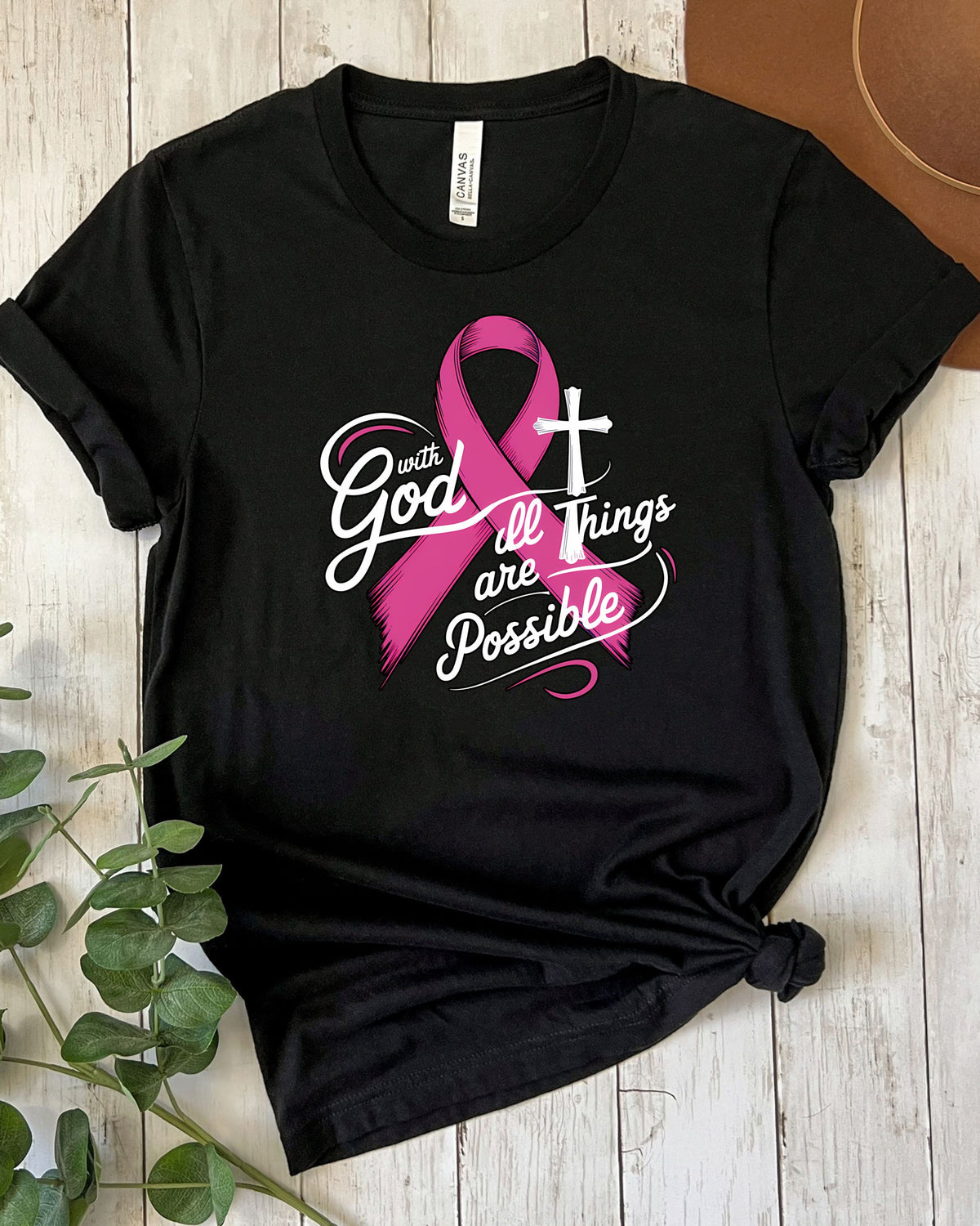 A black short-sleeve graphic T-shirt laid flat on a light wood surface, styled with a pink ribbon, white script text reading 'with God all things are possible,' and a pink-and-white cross design on the front.