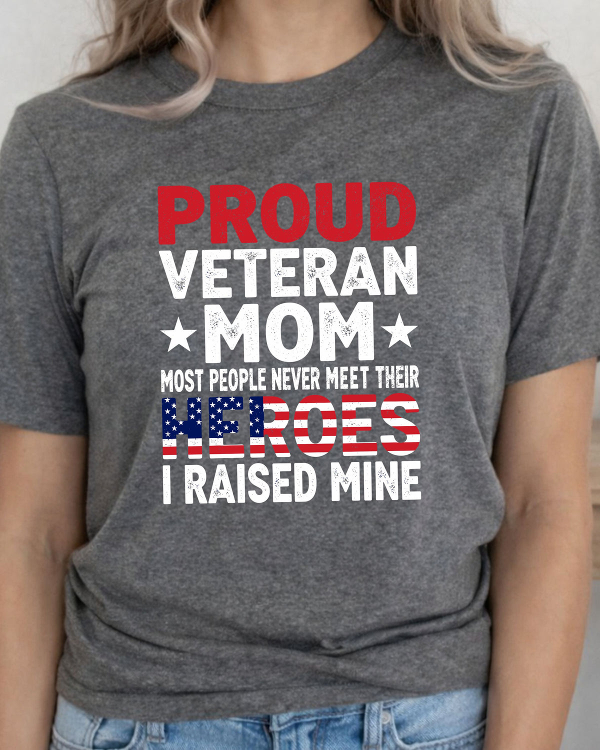 A woman wearing a heather gray graphic t-shirt with the text 'PROUD VETERAN MOM MOST PEOPLE NEVER MEET THEIR HEROES I RAISED MINE' in red, white, and blue patriotic colors.