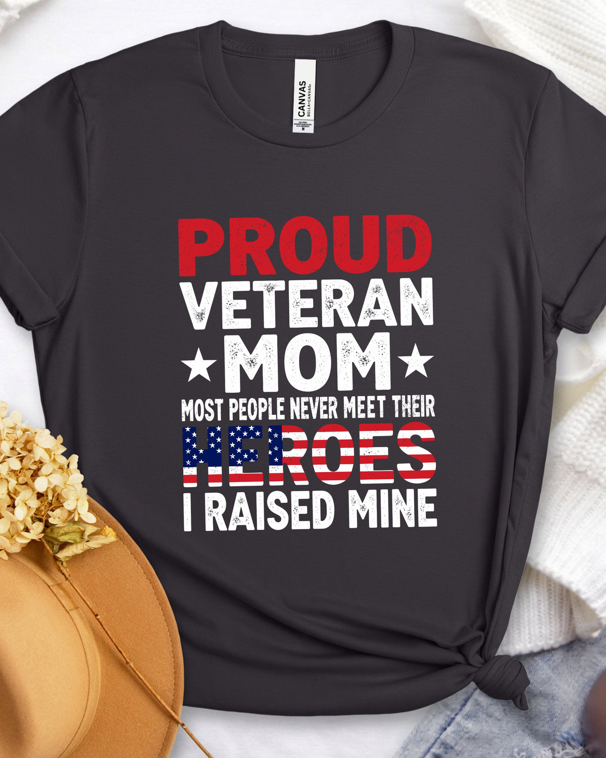 A black graphic t-shirt laid flat with a patriotic design that reads 'PROUD VETERAN MOM MOST PEOPLE NEVER MEET THEIR HEROES I RAISED MINE' in red, white, and blue. The shirt is styled with a tan hat, dried flowers, and a white knit blanket.