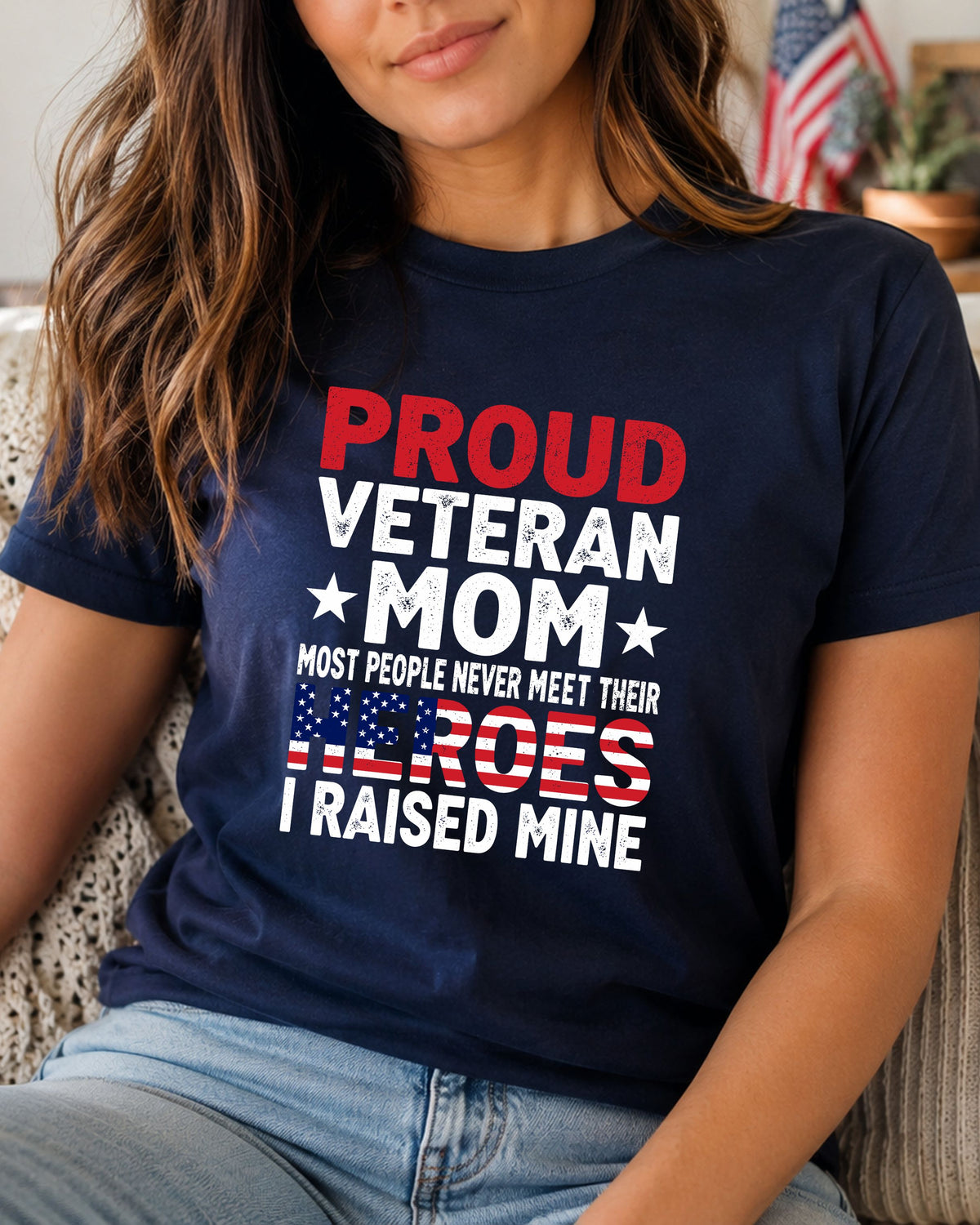 A woman wearing a navy blue graphic t-shirt with the text 'PROUD VETERAN MOM MOST PEOPLE NEVER MEET THEIR HEROES I RAISED MINE' in red and white, paired with light blue jeans.