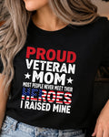 A woman wearing a black graphic t-shirt with the text 'PROUD VETERAN MOM MOST PEOPLE NEVER MEET THEIR HEROES I RAISED MINE' in red, white, and blue patriotic colors, paired with blue jeans.