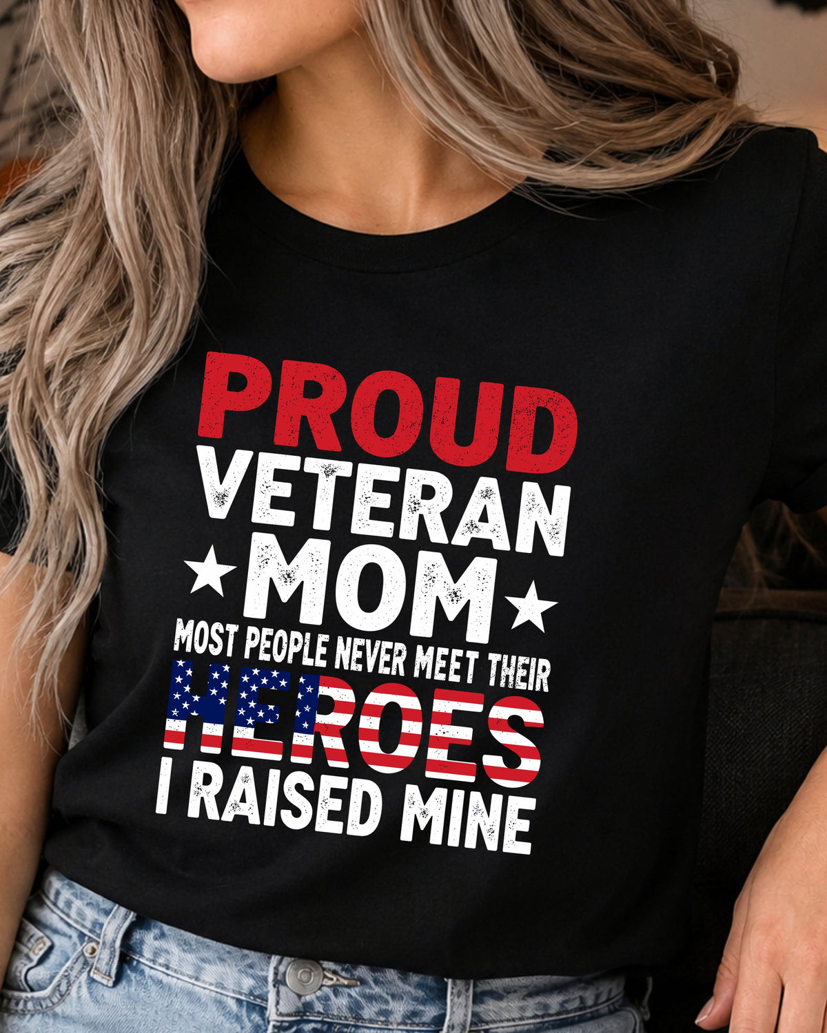 A woman wearing a black graphic t-shirt with the text 'PROUD VETERAN MOM MOST PEOPLE NEVER MEET THEIR HEROES I RAISED MINE' in red, white, and blue patriotic colors, paired with blue jeans.