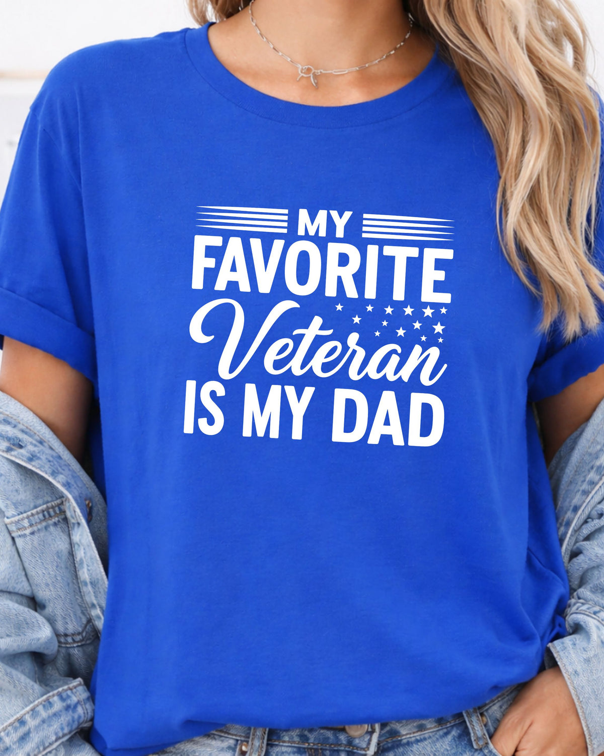 A woman wearing a bright blue short-sleeve t-shirt with the text 'MY FAVORITE Veteran IS MY DAD' printed in white across the front, styled with rolled-up denim jeans.