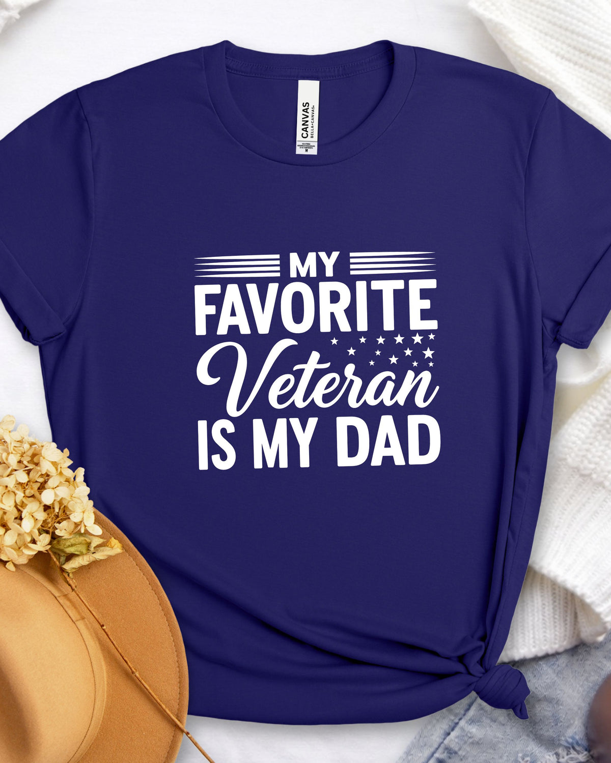 A navy blue graphic t-shirt with the text 'MY FAVORITE Veteran IS MY DAD' in white, styled in a flat-lay with a tan hat, white knit fabric, and denim on a white background.