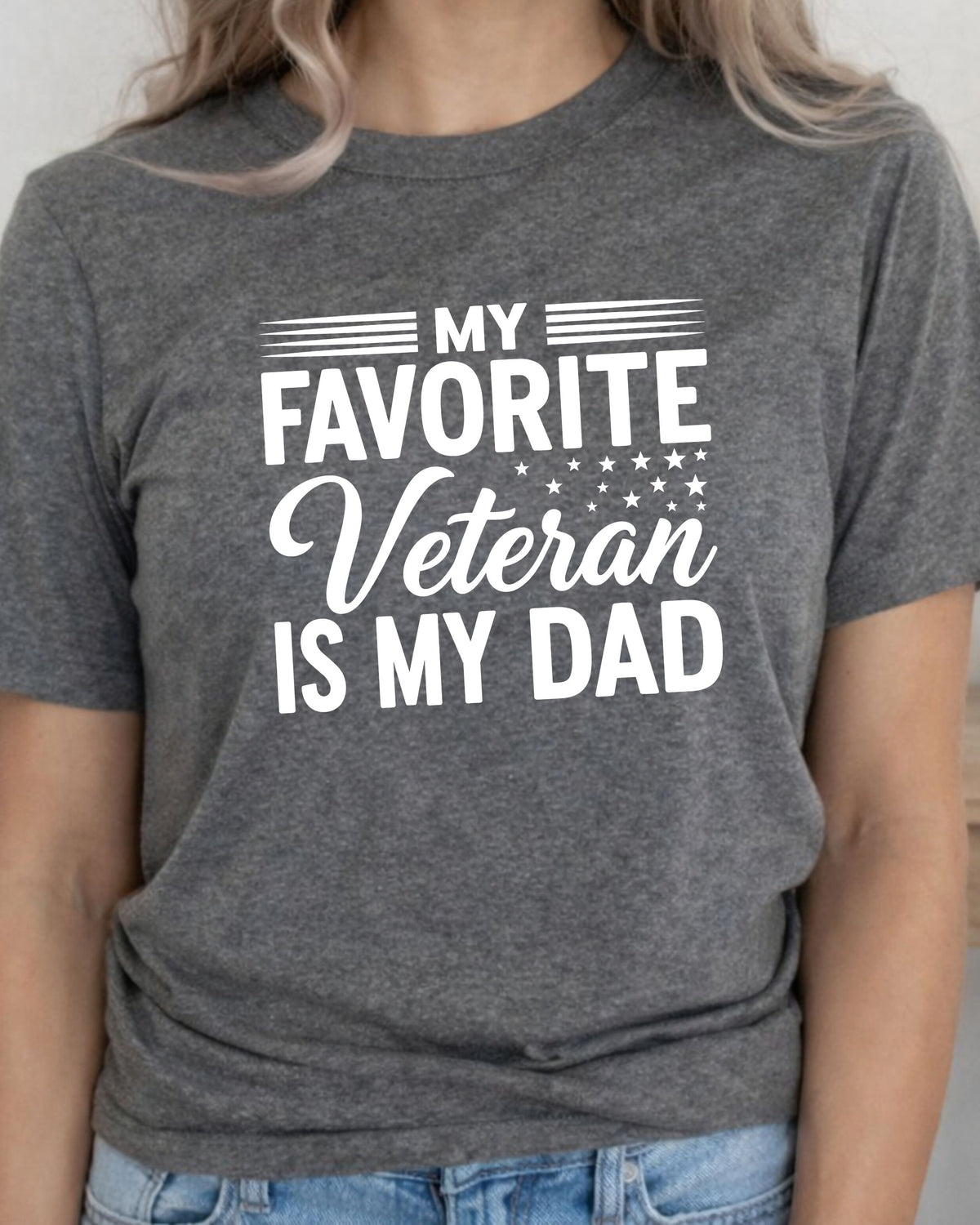 A woman wearing a heather gray t-shirt with a white graphic print that reads 'MY FAVORITE Veteran IS MY DAD' with stars and stripes details, paired with blue jeans.