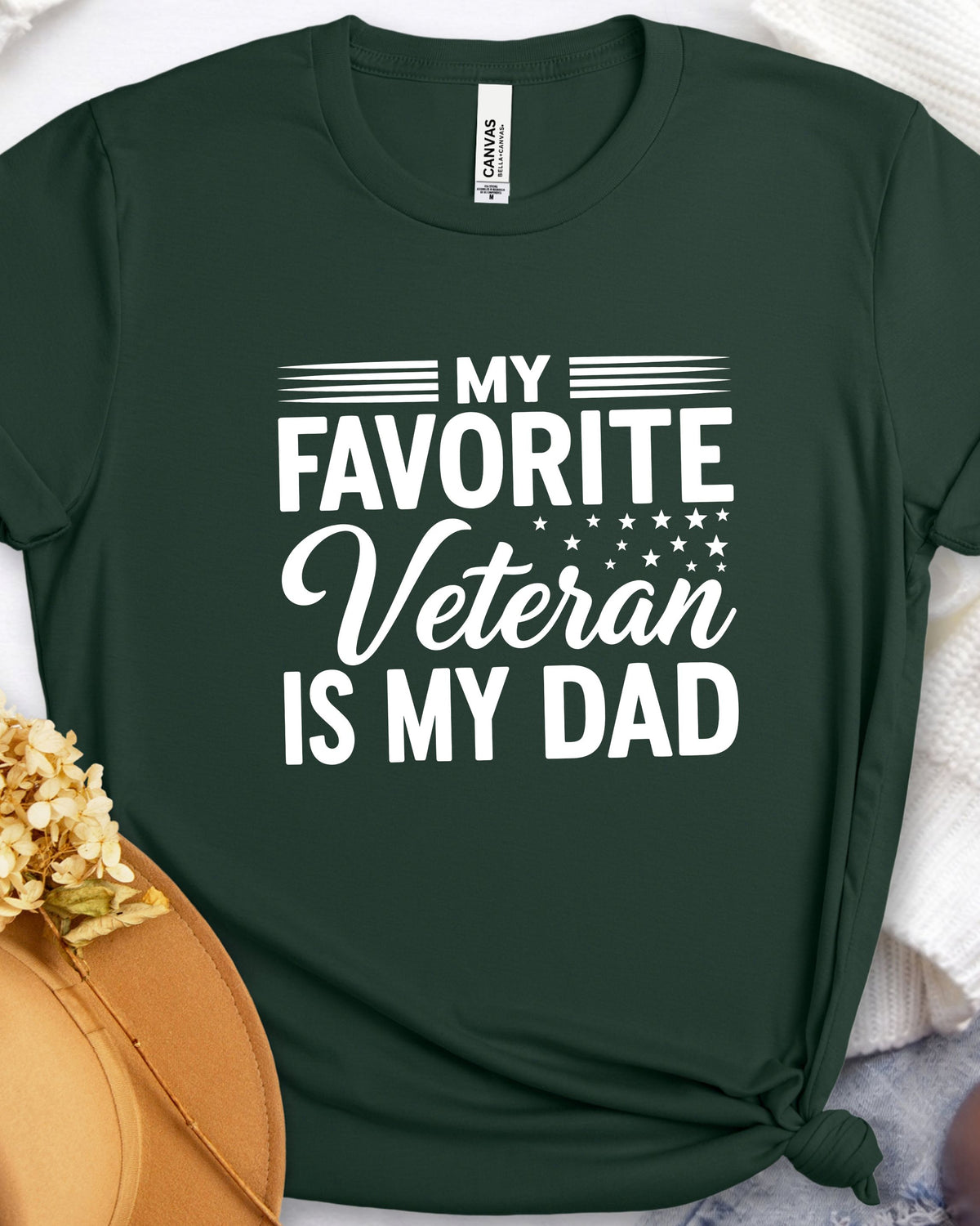 A dark green short-sleeve t-shirt with a white graphic print reading 'MY FAVORITE Veteran IS MY DAD' with stars and stripes details, styled flat-lay on a white textured blanket with a tan hat and dried flowers in the corner.