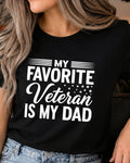 A woman wearing a black short-sleeve graphic T-shirt with the text 'MY FAVORITE Veteran IS MY DAD' in white, paired with light blue jeans.