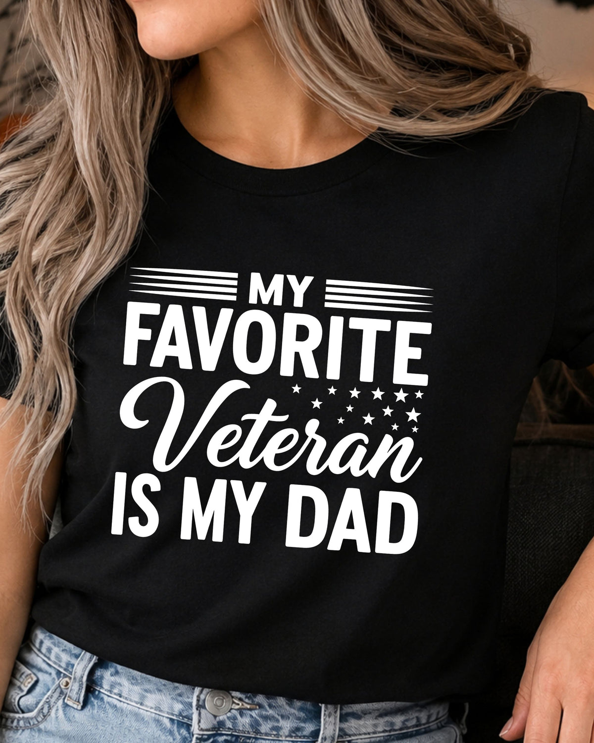 A woman wearing a black short-sleeve graphic T-shirt with the text 'MY FAVORITE Veteran IS MY DAD' in white, paired with light blue jeans.