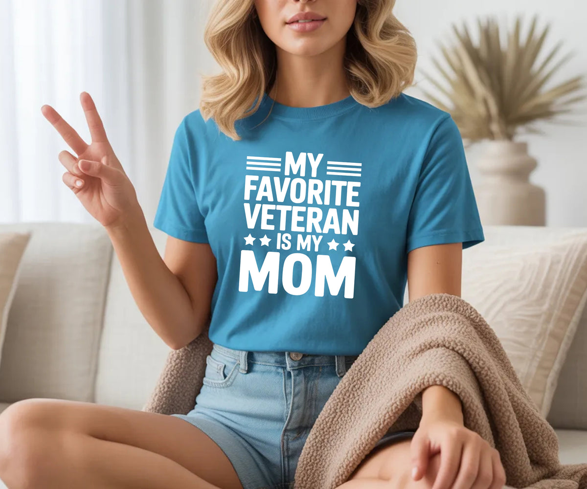 A woman sitting on a beige couch wearing a blue graphic t-shirt with the text 'MY FAVORITE VETERAN IS MY MOM' in white. She is making a peace sign with her hand, wearing light-wash denim shorts, and a brown knit blanket is draped over her lap.