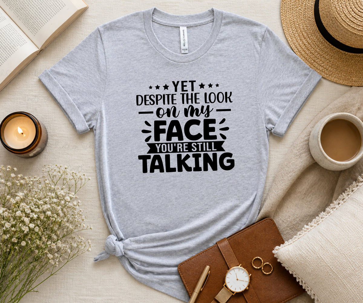 A light heather gray graphic t-shirt with black text reading 'YET DESPITE THE LOOK ON MY FACE YOU'RE STILL TALKING', styled in a cozy flat-lay with a straw hat, cup of coffee, candle, notebook, and flowers on a beige fabric background.