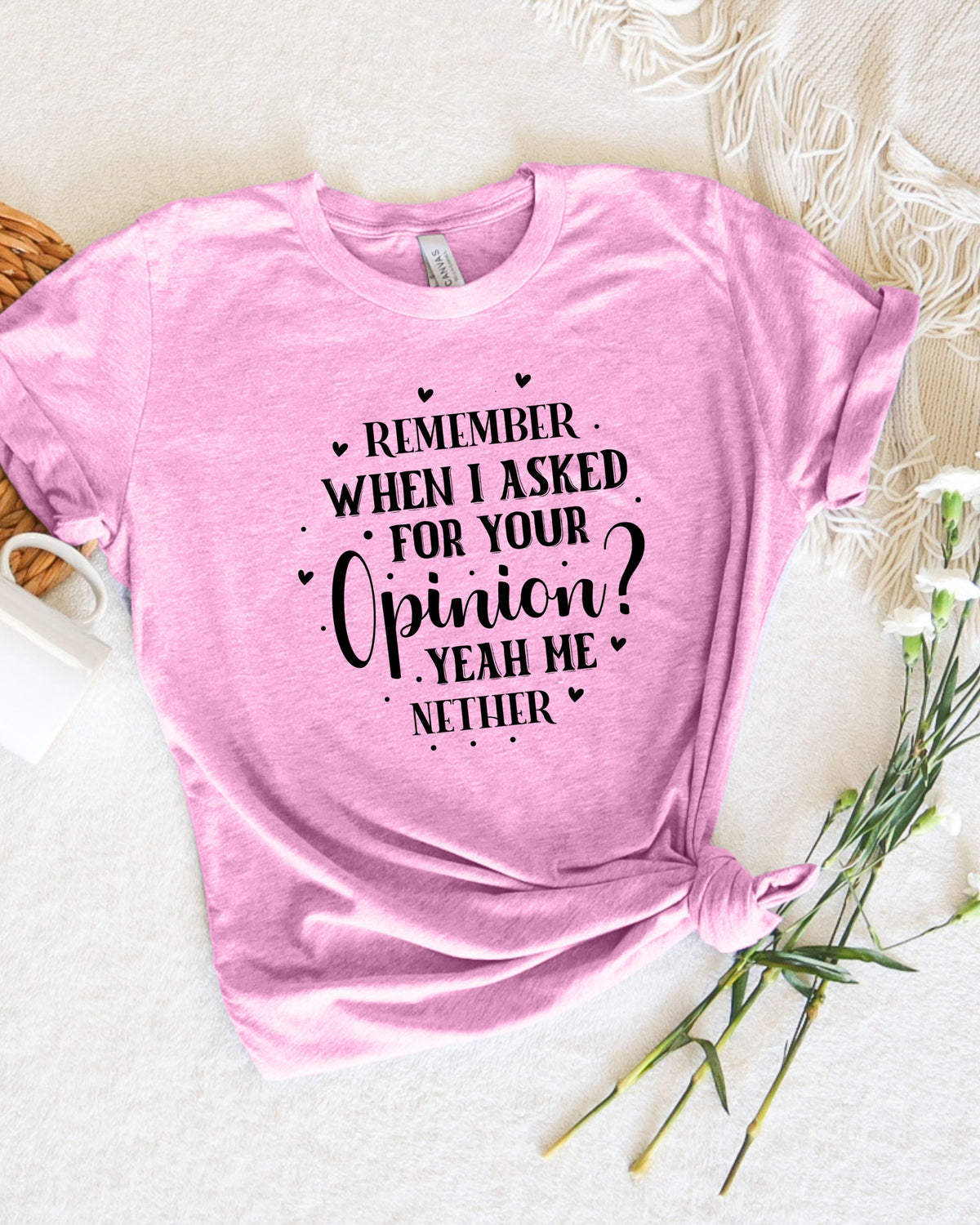 A pink short-sleeve graphic T-shirt laid flat on a light cream fabric background, styled with a small white mug, woven basket edge, and white flowers. The shirt has black text reading, 'REMEMBER WHEN I ASKED FOR YOUR Opinion? YEAH HE NETHER' with small heart and dot graphics.