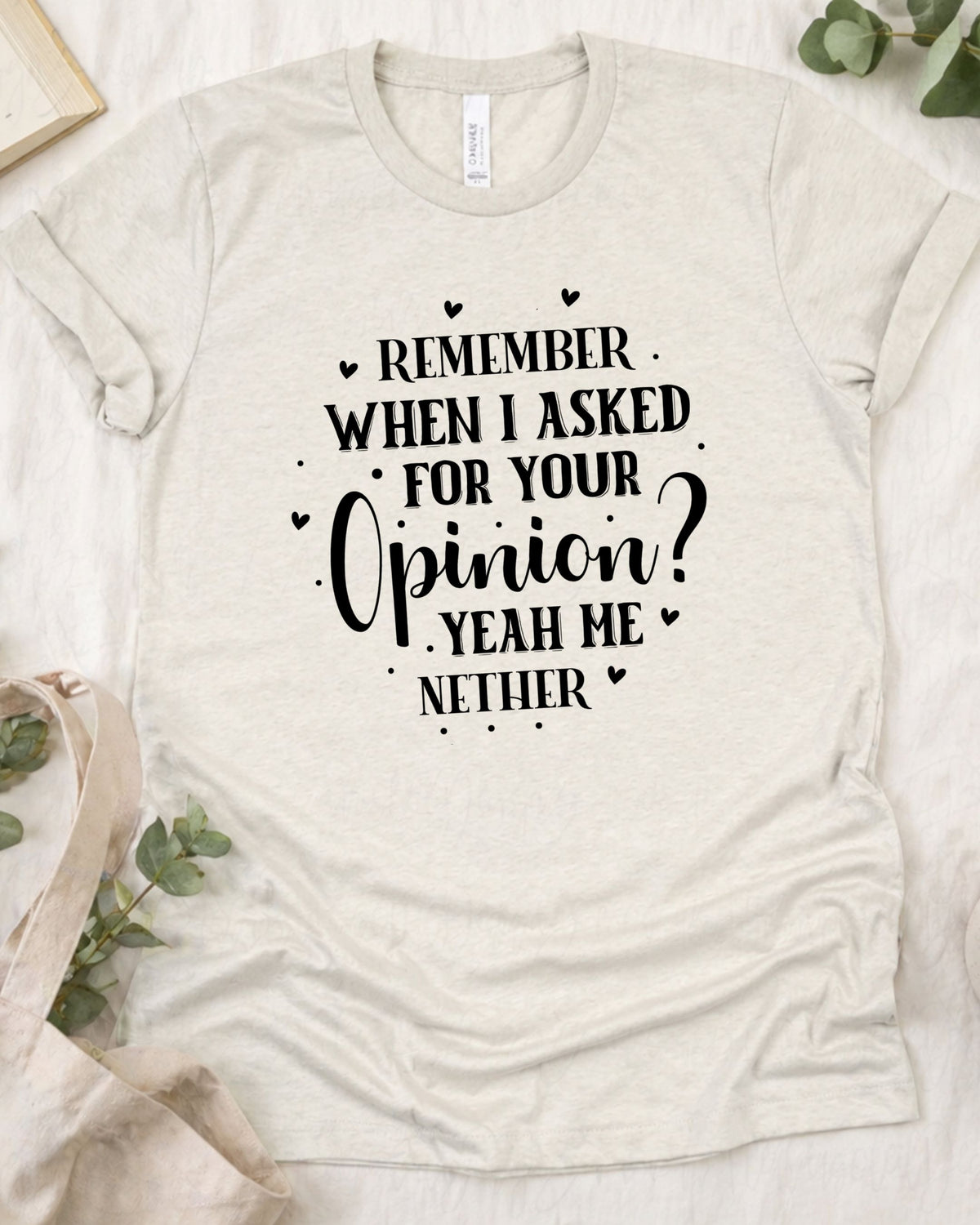 A light beige heathered t-shirt with a black printed graphic that reads 'REMEMBER WHEN I ASKED FOR YOUR Opinion? YEAH ME NETHER' surrounded by small heart doodles. The t-shirt is displayed in a flat-lay arrangement on a white textured fabric background with decorative eucalyptus leaves and a neutral fabric bag.