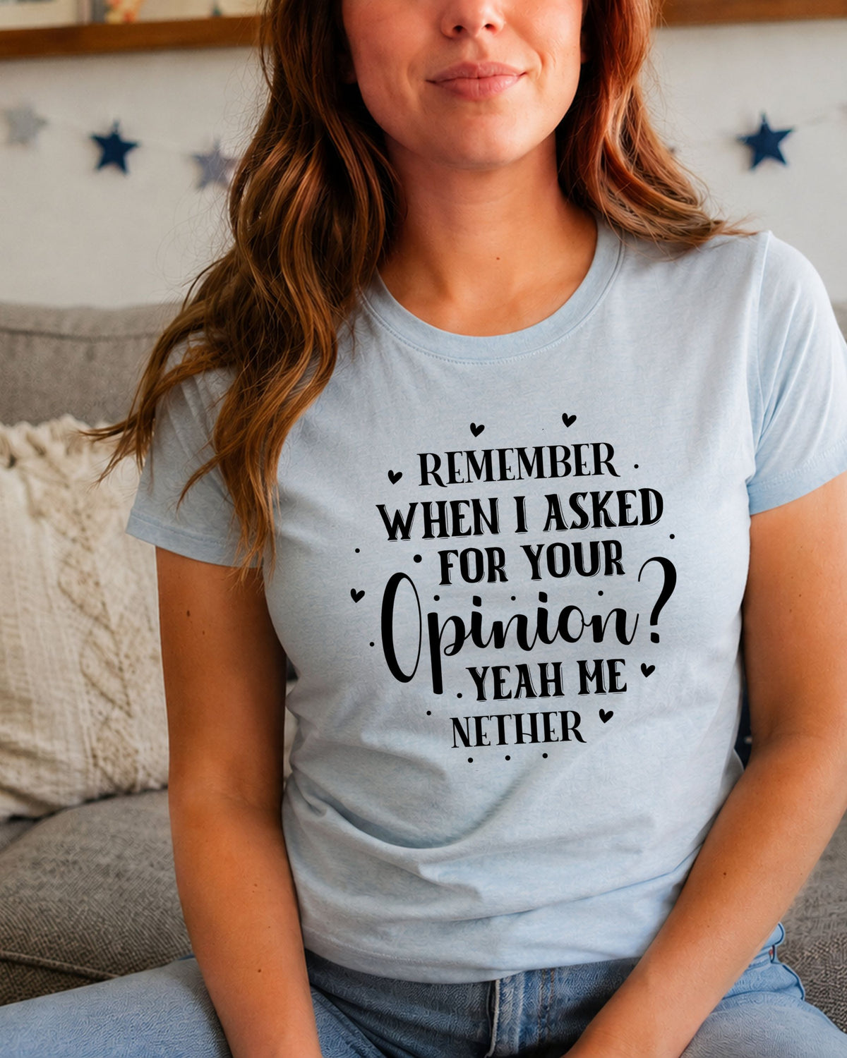 A woman wearing a light blue graphic t-shirt with black text that reads 'REMEMBER WHEN I ASKED FOR YOUR OPINION? YEAH HE NETHER' with small heart and dot accents, sitting on a couch in a living room setting.
