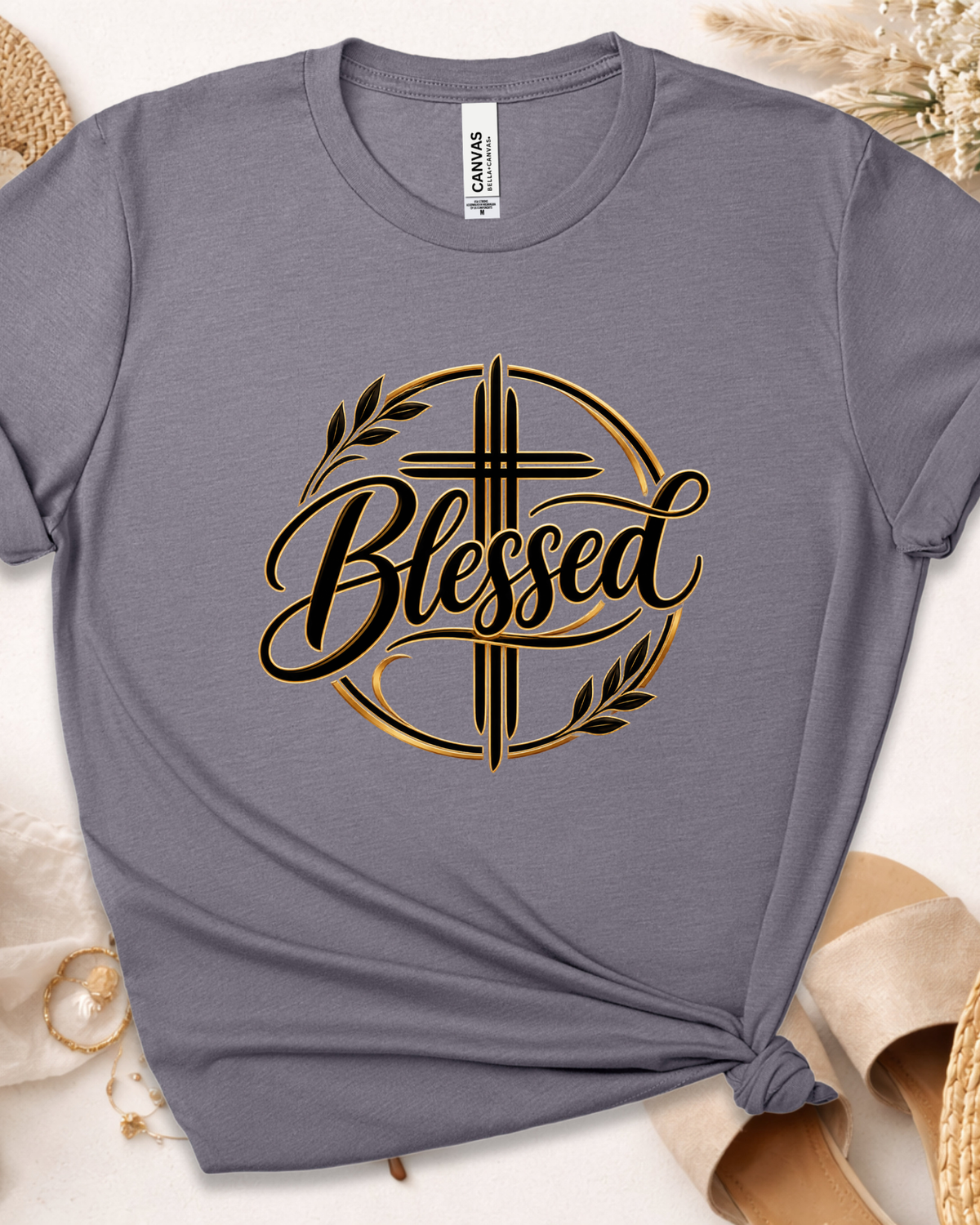 A heather gray short-sleeve t-shirt with a black and gold graphic on the chest featuring a cross, olive branches, and the word 'Blessed'. The shirt is styled in a flat-lay with neutral props including a straw hat, a beige shoe, and dried grass on a light beige surface.